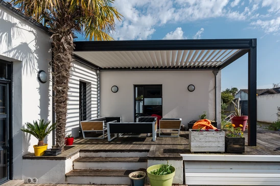 Terrasse couverte moderne avec pergola bioclimatique à lames orientables noires, mobilier d'extérieur gris contemporain, vue sur villa en pierre naturelle et végétation méditerranéenne